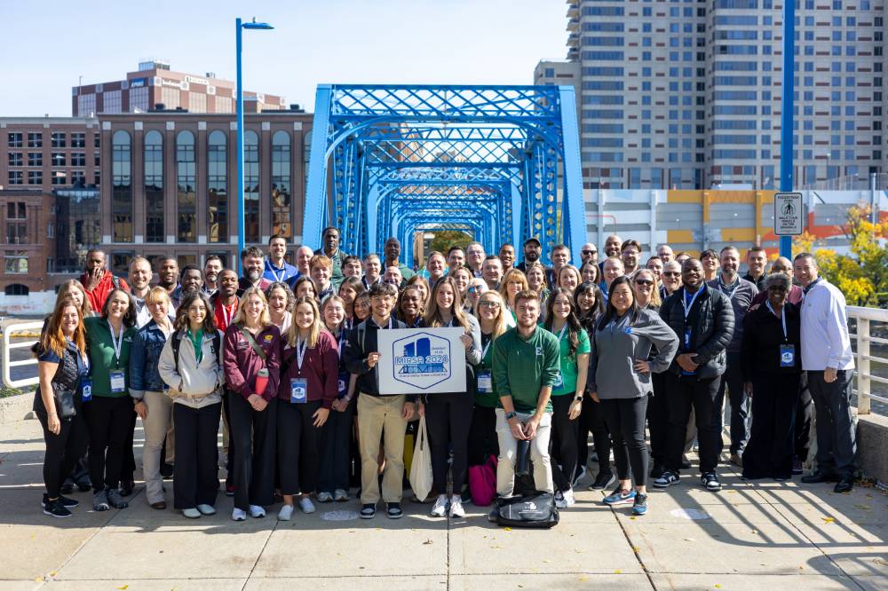 A large group of people standing outside in front of a blue bridge.